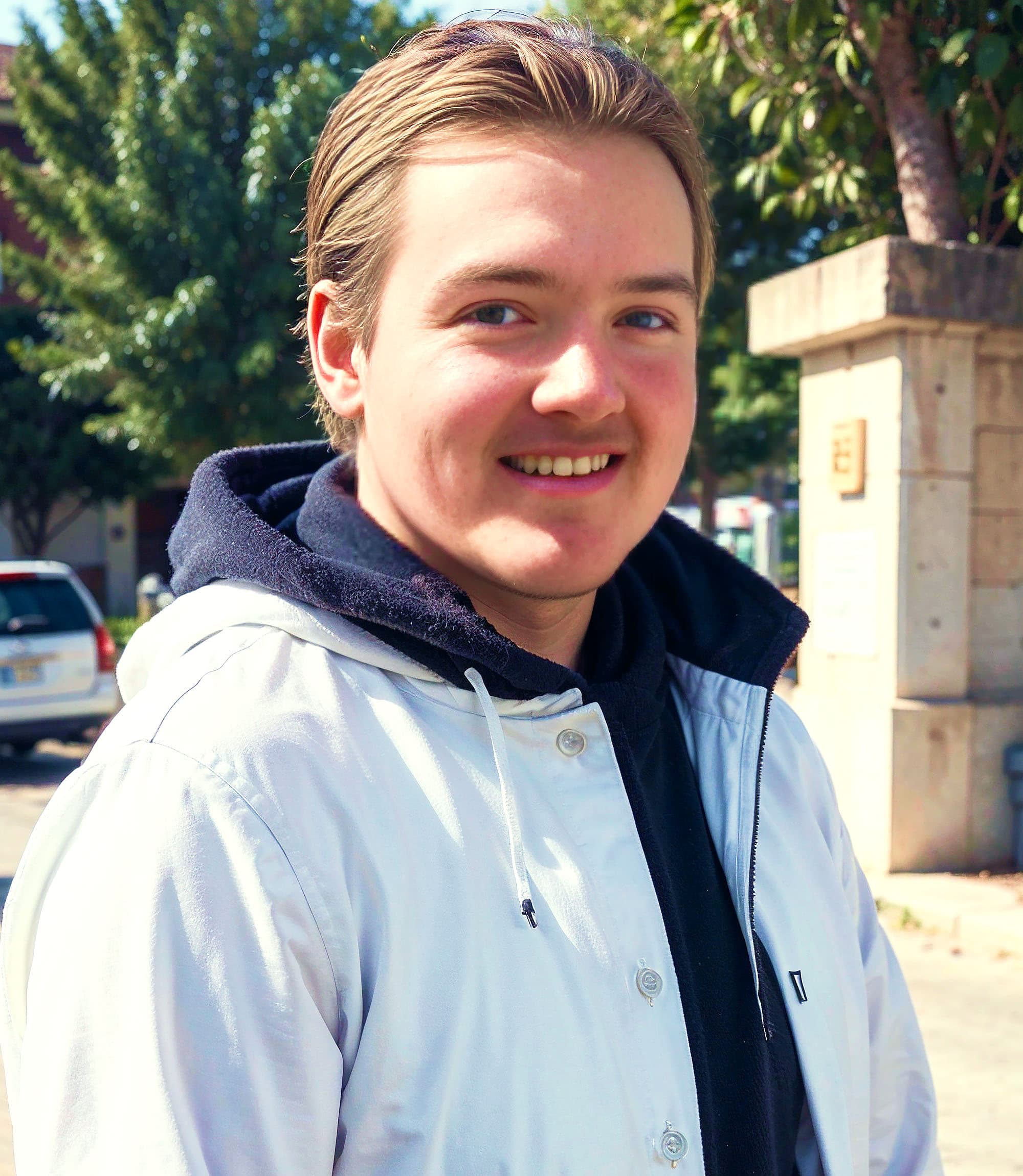 A young man stands outdoors. He is wearing a white coat.