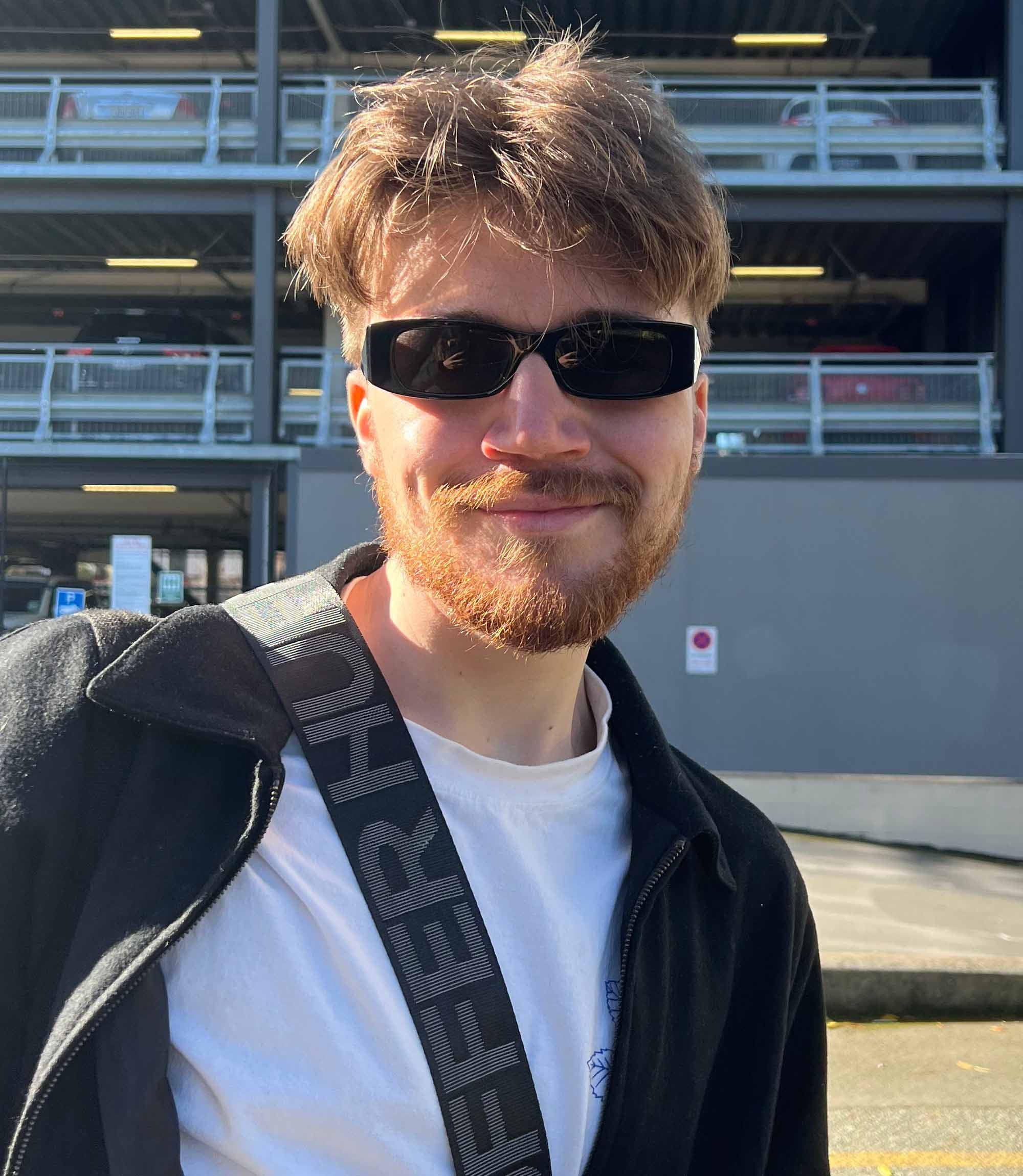 A young man with a light beard and mustache stands outdoors. He is wearing black sunglasses, a white t-shirt under a black zip jacket.